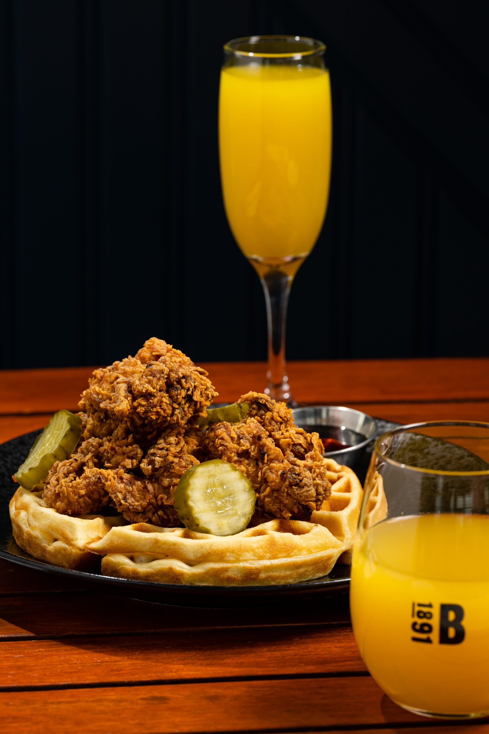 black dining plate with fried chicken and waffles with sliced pickles on the side