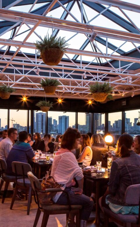 Rooftop restaurant dining room with guests, hanging plants, glass ceiling, and city skyline views at sunset.