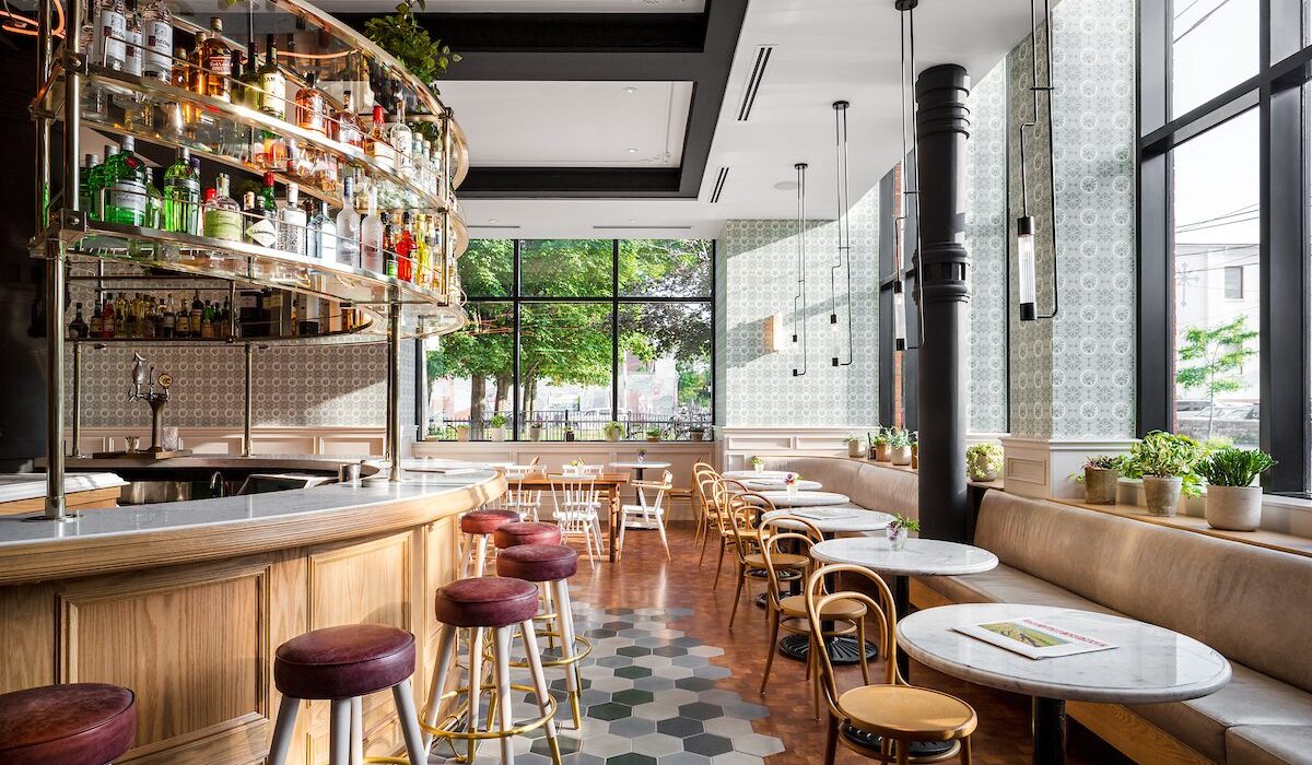 Sunlit cafe and bar area with large glass windows, dining tables, and indoor plants.