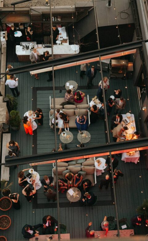 Overhead view of guests mingling on a rooftop patio with lounge seating, cocktail tables, bar service, and string lights.
