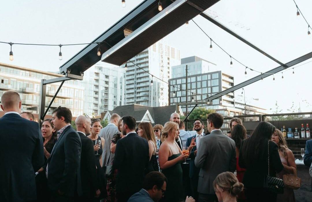 Crowd attending a cocktail reception on a heated outdoor patio surrounded by city buildings.