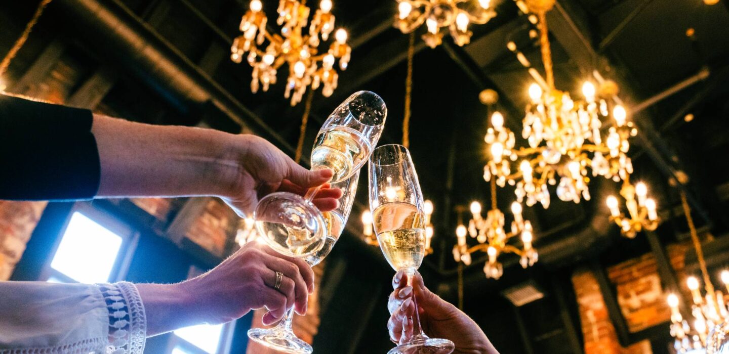 Close-up of hands clinking champagne glasses in a celebration under ornate chandeliers.
