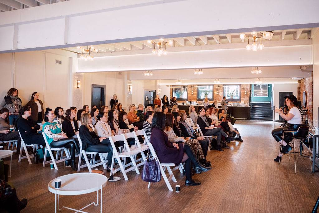 Speaker addressing a seated crowd during a seminar in an event space with exposed brick walls.