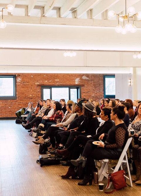 Crowd watching two speakers hosted on barstools in an event space with exposed brick walls.