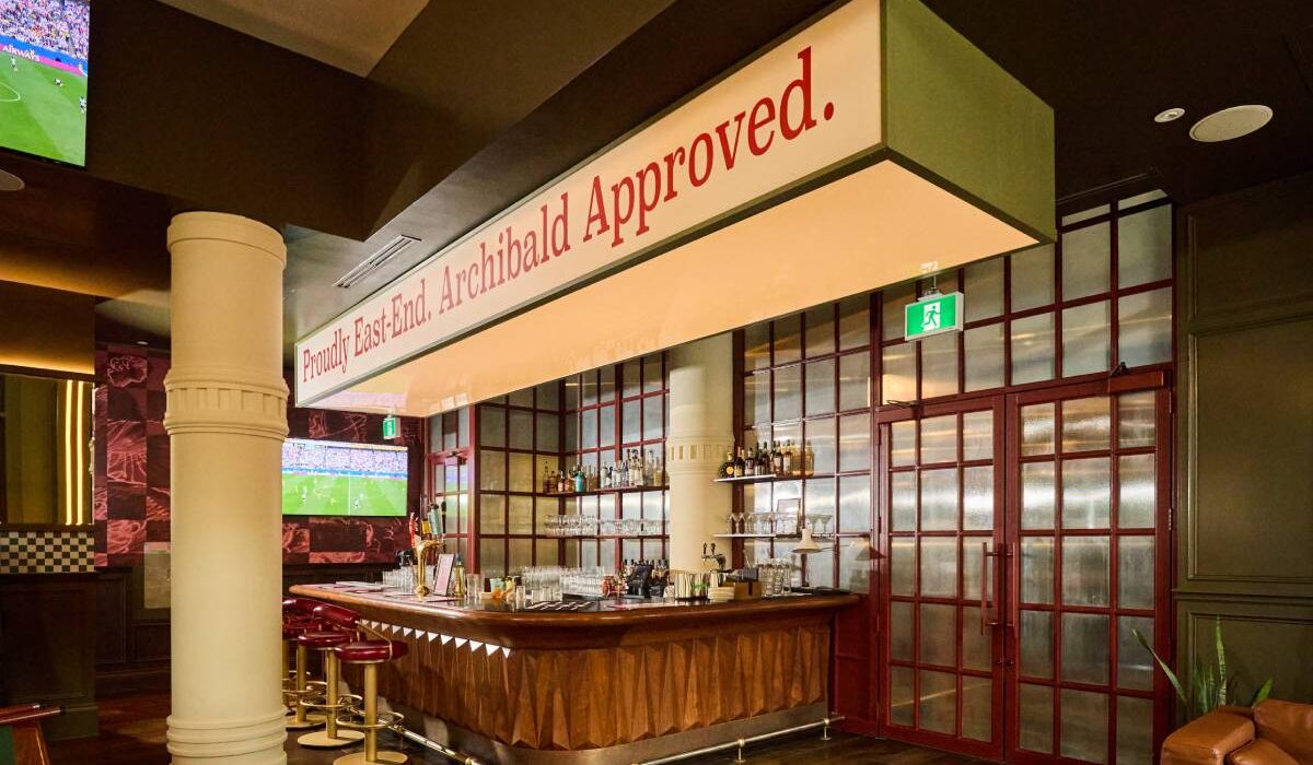 Restaurant bar with wood detailing, red-framed glass doors, bar seating, televisions, and an illuminated “Proudly East-End. Archibald Approved.” sign.