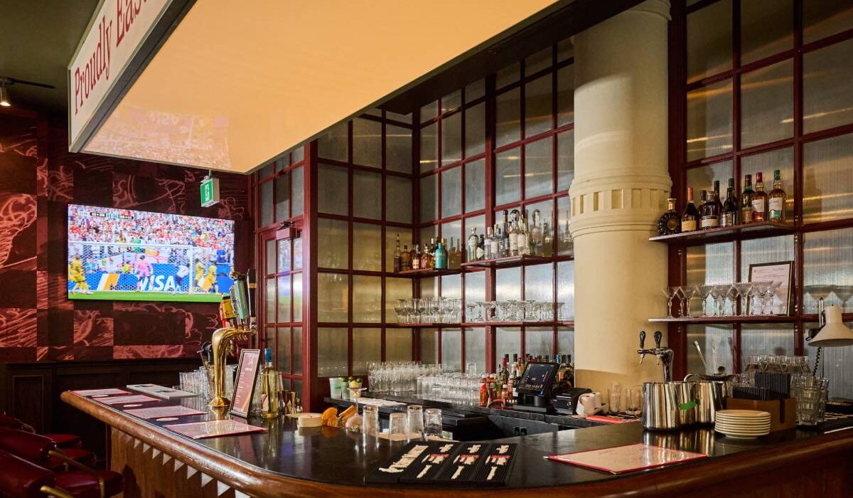 Restaurant bar with a curved wooden counter, red bar stools, liquor shelves, beer taps, and a television showing a soccer match.