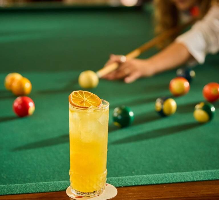 Yellow citrus cocktail garnished with a dried orange slice on a pool table, with a person playing pool in the background.